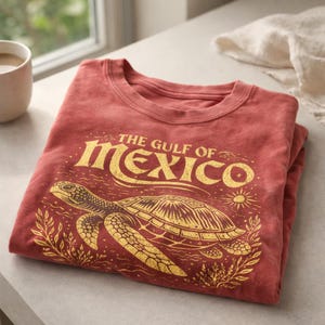 Pode incluir: Uma camiseta vermelha dobrada com um gráfico dourado de uma tartaruga marinha e o texto "THE GULF OF MEXICO". O design inclui um sol, ondas e folhagem. Uma caneca branca e um pano branco estão no fundo.