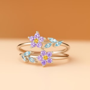 May include: A silver ring with two purple flower designs, each with a yellow centre, and light blue leaf accents. The ring has a double band design. The ring is set against a warm, neutral background.