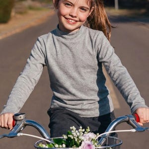 May include: A long-sleeved grey mock neck top worn by a child riding a bicycle with a basket of flowers. The text LittleCubbies Childrens Clothing is printed in green at the bottom of the image. The top features a simple design and fitted sleeves.