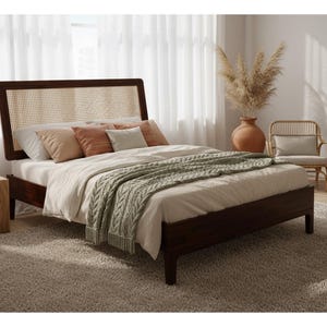 May include: A dark wood bed frame with a woven headboard, dressed with neutral-toned bedding and decorative pillows. A green knit throw blanket adds texture. The room features a neutral rug, a side table, and a woven chair, creating a cozy bedroom setting.