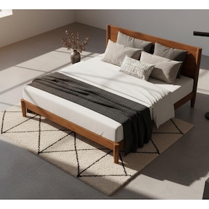 May include: A wooden bed frame with a white mattress, pillows, and a dark gray blanket. A cream-colored rug with a black geometric pattern is in front of the bed. A black floor lamp is to the right of the bed. A vase with dried flowers is on the left.
