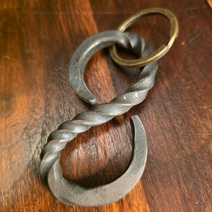 May include: A hand-forged iron keychain featuring an S-shaped metal piece with a twisted texture throughout its center. The dark gray metal component is attached to a circular brass-toned split ring, creating a functional and durable accessory.