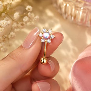 Può includere: Un piercing per ombelico color oro con un motivo floreale. Presenta una pietra centrale iridescente e cristalli trasparenti. Il bilanciere curvo ha una piccola sfera dorata in basso.