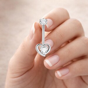 May include: A silver belly button ring with a heart-shaped design. The ring features a large, clear, round gemstone at the top and a heart-shaped gemstone at the bottom. The heart is outlined with a silver frame.