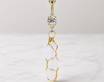 Elegante joya dorada para piercing de ombligo con gema transparente y colgante de cristal curativo envuelto en alambre sobre una elegante superficie de piedra.