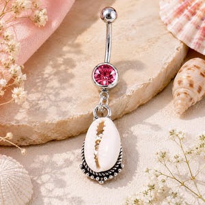 May include: A silver-toned navel bar featuring a curved barbell with a round, faceted pink crystal at the top and a dangling white cowrie shell charm set in a decorative, oxidised silver-toned metal frame at the bottom.