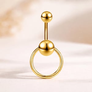 Puede incluir: Un piercing para el ombligo de color dorado. La joya presenta una pequeña bola en la parte superior, una barra recta, una bola más grande en el centro y un anillo circular en la parte inferior. El metal tiene una superficie brillante y reflectante.