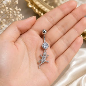 Peut inclure: Un piercing de nombril argenté composé d'une barre incurvée avec une pierre précieuse ronde et transparente sur le dessus, ainsi qu'un pendentif en forme de lézard incrusté de petits cristaux transparents. Le métal est poli et brillant.
