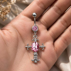 May include: A silver-colored belly button ring featuring a cross design. The ring has a small, round gem at the top and a larger, oval pink gem at the center of the cross. The cross has decorative flourishes on the arms and base.