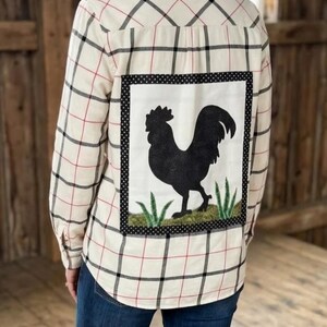 May include: A long-sleeve button-down shirt in a cream, black, and red plaid pattern. The back features a large, rectangular white patch with a black polka-dot border, showcasing a black silhouette of a rooster standing on green grass.
