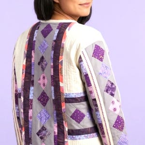 May include: Cream-colored quilted jacket with a patchwork design featuring purple, gray, and brown diamond shapes. The jacket has a unique, handcrafted aesthetic, with a mix of patterned fabrics and decorative stitching. The overall style is a blend of comfort and artistic expression.