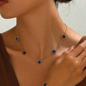 Può includere: Un set di gioielli color oro con collana, bracciale e orecchini. Ogni pezzo presenta un design a forma di trifoglio con un centro blu scuro. La collana e il bracciale hanno più ciondoli a trifoglio, mentre gli orecchini ne hanno uno ciascuno.