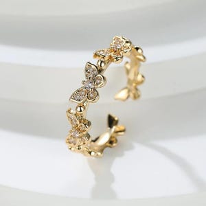 Könnte beinhalten: Ein goldfarbener Ring mit Schmetterlingsmotiv. Der offene Ring ist mit kleinen, funkelnden Edelsteinen verziert. Die Schmetterlinge sind um das Band angeordnet und erzeugen einen zarten und eleganten Look. Der Ring ist ein Schmuckstück.