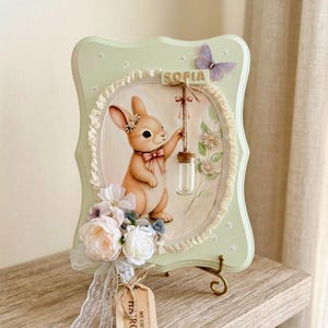 May include: A light green scalloped wooden plaque featuring an oval illustration of a brown rabbit holding a small glass vial. The plaque is decorated with white fabric flowers, a purple butterfly, and a wooden tag labeled MY FIRST HAIRCUT.