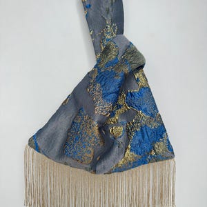 May include: A knot-handle bag crafted from a grey fabric featuring a textured pattern of metallic gold and vibrant blue abstract shapes. The bottom edge of the bag is finished with a long, delicate fringe in a shimmering gold tone.
