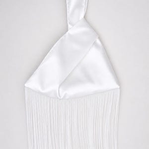 Op de afbeelding: Een witte satijnen clutch met een geknoopt handvat en lange franjes. De tas heeft een driehoekige vorm en is gemaakt van een gladde, glanzende stof. De franjes hangen van de onderkant van de tas.