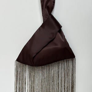 May include: A dark brown satin handbag with a knotted handle. The bottom edge features long, shimmering fringe in shades of gold and brown. The bag is displayed against a plain white background, highlighting its elegant design.
