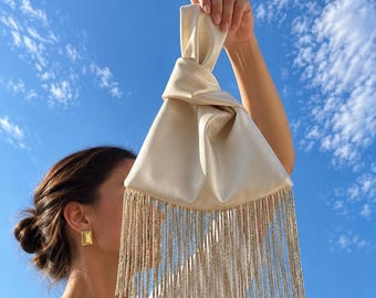 May include: A cream-colored satin handbag featuring a knotted handle design and a long, shimmering fringe trim that hangs from the bottom edge. The fabric has a smooth, reflective finish, and the fringe consists of numerous thin, metallic strands.