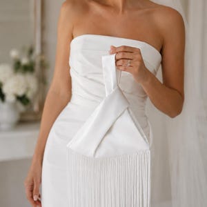 Peut inclure: Pochette en satin blanc avec franges. La pochette est tenue par une personne portant une robe bustier blanche. L'image est prise à l'intérieur avec un éclairage doux, suggérant une occasion de mariage ou formelle.
