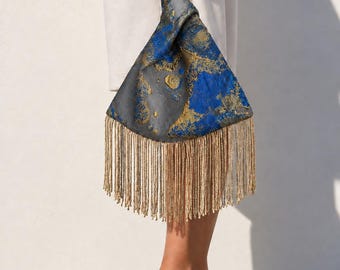 Puede incluir: Un bolso de mano triangular de tela con un patrón abstracto en gris oscuro y azul vibrante con detalles metálicos dorados. El bolso tiene un asa superior anudada y está rematado con un fleco largo y denso de hilos dorados en el borde inferior.