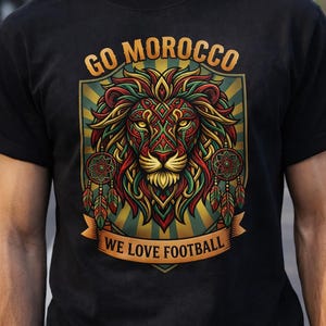 Puede incluir: Camiseta negra con un diseño de león colorido. El diseño incluye el texto "GO MOROCCO" encima del león y "WE LOVE FOOTBALL" debajo. El león es de color rojo, verde y dorado, con atrapasueños a cada lado.