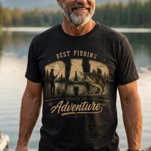 May include: A charcoal grey short-sleeve t-shirt featuring a graphic print with the text "BEST FISHING DAD" and "HOOKED ON ADVENTURE". The design includes an illustration of a person fishing by a lake with a large fish jumping out of the water.