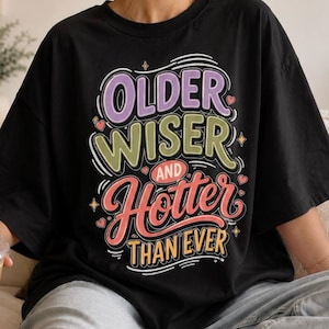 May include: Black t-shirt with the text "OLDER WISER AND HOTTER THAN EVER" in a colorful, playful design. The text is in a mix of fonts and colors, with hearts and stars. A casual, comfortable style.