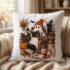 May include: A white square throw pillow featuring a colorful illustration of a ghost wearing an orange beanie, reading a book titled Ghost Stories while sitting in a chair. The scene includes black cats, pumpkins, autumn leaves, lanterns, and stacks of books.