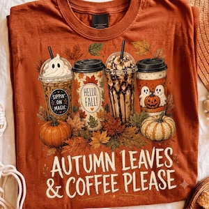 May include: A burnt orange t-shirt featuring a graphic of four coffee cups surrounded by autumn leaves and pumpkins. The cups display text including Sippin on Magic and Hello Fall. The bottom of the shirt features the text Autumn Leaves & Coffee Please.