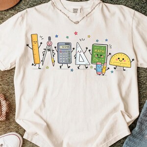 May include: An off-white short-sleeve t-shirt featuring a graphic print of anthropomorphic school supplies including a ruler, compass, calculator, triangle, a green chalkboard labelled MATH, a pencil, and a protractor, all with smiling faces.