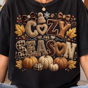 May include: A black t-shirt featuring a graphic print with the text "COZY SEASON" in large, textured letters. The design includes brown bows, a knit beanie, maple leaves, acorns, and five pumpkins with various patterns like plaid and cable knit.
