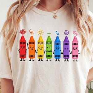 Peut inclure: Un t-shirt couleur crème arborant un motif de sept crayons de couleur animés alignés. Chaque crayon présente une expression faciale unique et un symbole au-dessus, incluant un gribouillis, une spirale, un soleil, un signe moins, une goutte, une spirale et des cœurs.