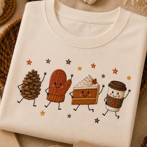 Op de afbeelding: Een crèmekleurig t-shirt met een grafische print van vier antropomorfe figuren: een dennenappel, een want, een stuk taart en een koffiebeker, allemaal omringd door kleine kleurrijke sterretjes op een effen stof.