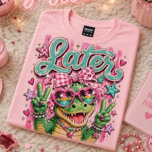 May include: A light pink t-shirt featuring a graphic of a green alligator wearing heart-shaped sunglasses with a sunset palm tree print and a pink gingham bow. The alligator is making peace signs with both hands. The word Later is printed above.