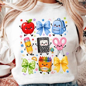 May include: A white sweatshirt featuring a colorful graphic print of anthropomorphic school supplies including a red apple, blue bow, blue backpack, yellow pencil, black and white composition notebook, pink scissors, green bow, orange crayon box, and yellow bow.