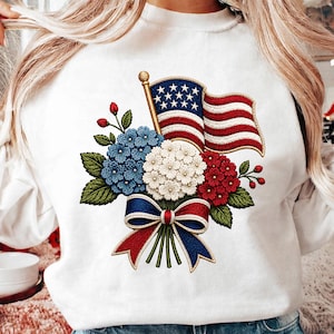 May include: A white long-sleeve sweatshirt featuring a central graphic of a bouquet of blue, white, and red hydrangea flowers tied with a matching patriotic ribbon, accompanied by a waving American flag on a gold pole with a ball finial.