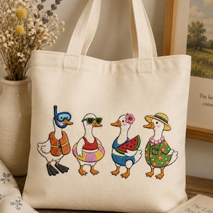 May include: A cream-colored canvas tote bag featuring an embroidered design of four cartoon ducks. Each duck is dressed in summer attire, including a life vest, sunglasses, a swim ring, and a Hawaiian shirt. The bag has two handles.