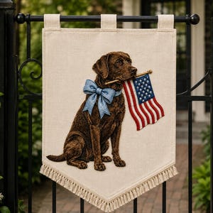 May include: A rectangular off-white fabric banner featuring an embroidered brown Labrador retriever sitting and holding a small American flag in its mouth. The dog wears a light blue bow around its neck. The banner has a fringed bottom edge.