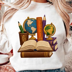 Puede incluir: Una sudadera blanca con un gráfico bordado de material escolar que incluye un libro abierto sobre un libro cerrado, dos globos terráqueos, una regla, un lápiz morado y un lápiz verde en un soporte marrón con un diseño de hoja al frente.