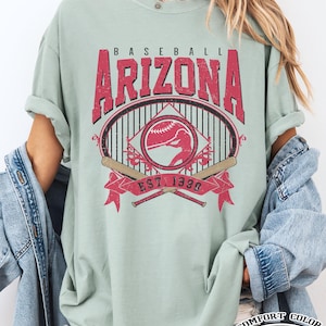 May include: A light green Comfort Color t-shirt with a distressed red graphic that says "Baseball Arizona" with a baseball and bats. The shirt also has the text "Est. 1998" and "Bay" at the bottom.