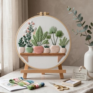 May include: A round embroidery hoop on a wooden stand featuring a stitched design of six potted plants on a shelf. The kit includes colorful embroidery floss skeins, a small box of needles, and a tag labeled Window-sill Garden Embroidery Kit.