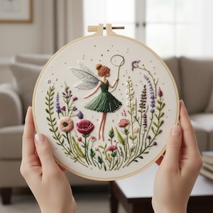 K&ouml;nnte beinhalten: Ein runder Stickrahmen mit einem Feen-Design. Die Fee, mit braunen Haaren und einem gr&uuml;nen Kleid, ist von bunten gestickten Blumen und Gr&uuml;n umgeben. Das Kunstwerk ist auf einem cremefarbenen Stoff angefertigt.