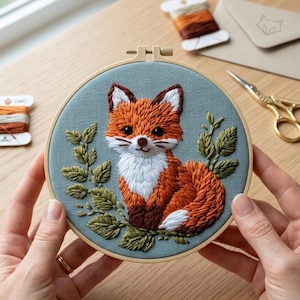 May include: An embroidered fox hoop art. The fox is rendered in shades of orange, brown, and white, with a detailed stitch pattern. The background is a muted teal fabric, and the design includes green leaves. The embroidery hoop is held by hands.