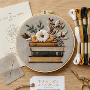Könnte beinhalten: Ein Stickrahmen mit einem gestickten Motiv aus einem Stapel von vier Büchern, gekrönt von einem Blumenarrangement aus weißen und gelben Blüten sowie grünen Blättern. Enthält vier Stickgarnstränge, eine Nadel und Anleitungskarten.