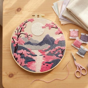 Könnte beinhalten: Ein fertiger Stickrahmen mit einer Landschaftsszene mit Fluss, Bergen und Kirschblütenbäumen. Das Kunstwerk verwendet rosa, graue und cremefarbene Fäden. Stickzubehör, einschließlich Garnrollen und Scheren, sind auf einer Holzoberfläche angeordnet.