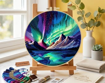 Könnte beinhalten: Ein Stickrahmen mit einer farbenfrohen Darstellung der Aurora Borealis, ausgestellt auf einer hölzernen Staffelei. Das Kunstwerk zeigt lebendige Farbtöne von Lila, Grün und Blau. Stickgarn, Nadeln und eine Anleitung sind ebenfalls sichtbar.
