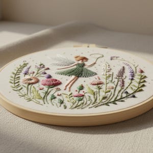 K&ouml;nnte beinhalten: Gesticktes Feen-Hoop-Art. Eine Fee in einem gr&uuml;nen Kleid und mit wei&szlig;en Fl&uuml;geln h&auml;lt einen Kreis, umgeben von bunten Blumen und Gr&uuml;n. Die Stickerei ist in einem Holzreifen gefasst.