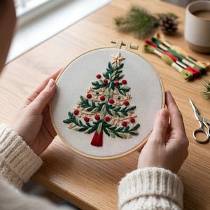 May include: A circular wooden embroidery hoop containing a white fabric panel with a hand-stitched Christmas tree design. The tree features dark and light green foliage, small red circular ornaments, gold thread accents, and a red tassel base.
