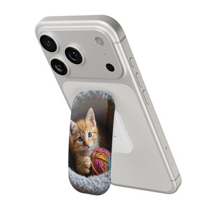May include: A phone grip featuring a ginger kitten with blue eyes nestled in a gray bed, holding a ball of yarn. The grip is attached to a silver smartphone.