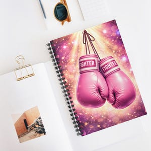 May include: A spiral-bound notebook featuring a vibrant illustration of pink boxing gloves. The gloves are suspended against a background of sparkling lights and rays. The word "FIGHTER" is printed on the wristbands. A gold paper clip and sunglasses are also visible.